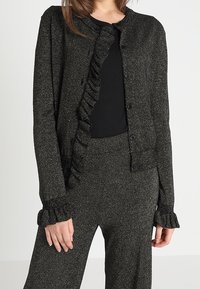 Zwarte, glinsterende cardigan met gerimpelde voorkant, ronde hals en knoopsluiting. Voorzien van gerimpelde manchetten, gecombineerd met een zwarte top en bijpassende broek.
