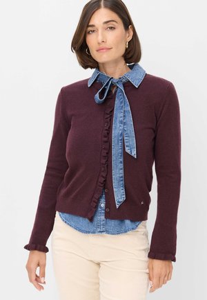 Kvinde iført en burgunderfarvet cardigan med flæsekant over en blå denimskjorte bundet ved halsen og lyse beige bukser, stående mod en hvid baggrund.
