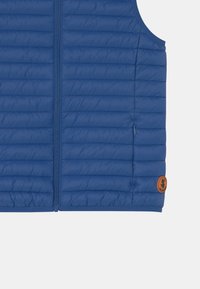 Blauer Steppweste mit gequilteter Struktur, durchgehendem Reißverschluss und einer kleinen Seitentasche. Mit einem runden braunen Logoakzent am Saum.