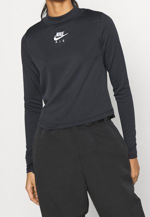 Personne portant un haut noir Nike Air côtelé à manches longues et un pantalon noir ample, debout devant un fond clair uni.