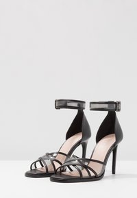 Svarta högklackade sandaler med genomskinliga detaljer, öppet tå-design, korsande remmar och fotledsrem med spänne, med en elegant finish.
