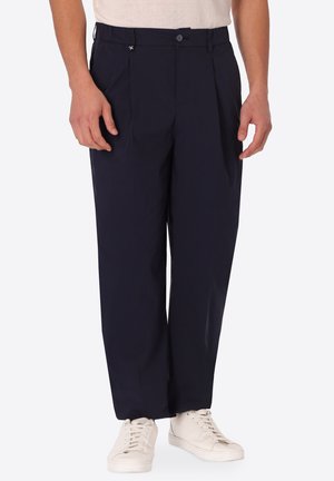 Uomo che indossa pantaloni plissettati blu navy e sneakers bianche, in piedi con le mani rilassate ai lati, visibile dalla vita ai piedi.