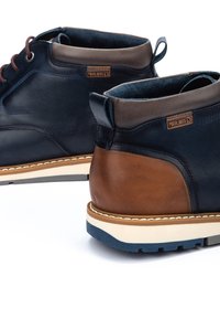 Dark blue leather ankle boots feature brown leather accents, beige sole, and round laces. Pikolinos branding visible on the side.