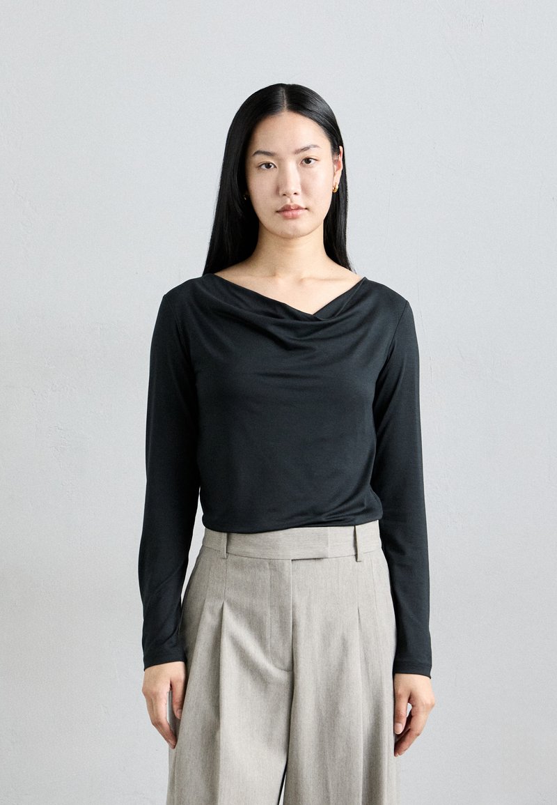 Black long-sleeve top with a draped neckline, paired with light grey pleated trousers. Smooth fabric with a fitted silhouette.