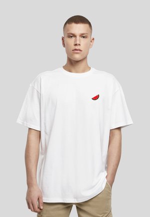 Jeune homme aux cheveux courts portant un t-shirt blanc oversize avec un petit graphique représentant une tranche de pastèque sur la poitrine et un pantalon beige.