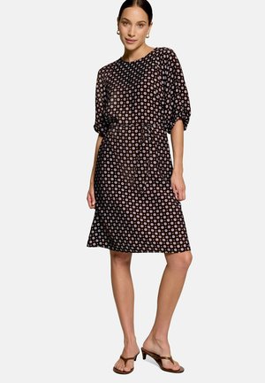 Femme portant une robe noire longueur genoux avec des motifs circulaires blancs, des manches bouffantes et une ceinture en tissu, associée à des sandales marron à talons.