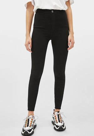Personne portant un jean skinny noir taille haute et des baskets blanches épaisses avec des accents orange, noir et bleu sarcelle, debout devant un fond uni.