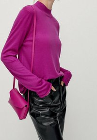 Een persoon draagt een fel magenta langemouwen top en een bijpassende schoudertas, gecombineerd met hooggeknoopte zwarte leren broek, met de handen in de zakken.