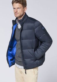 Marineblaue Pufferjacke mit horizontalem Steppmuster, blauer Innenverkleidung, hohem Kragen und Reißverschluss. Verfügt über ein kleines Logo am Ärmel.
