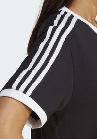 Černé sportovní tričko s bílými třemi pruhy na rukávech a bílým žebrovaným límcem, vyrobené z měkké látky.