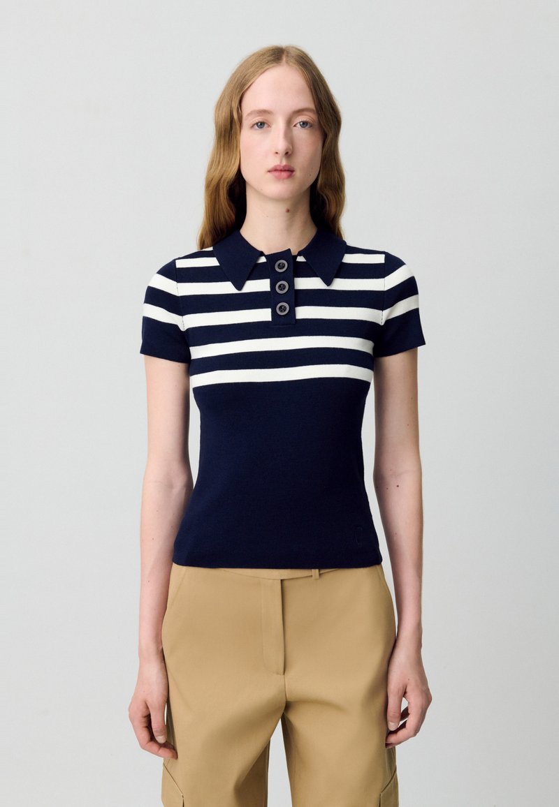 Polo a righe navy e bianche in un design aderente. Presenta un colletto e una chiusura con tre bottoni. Tessuto in maglia morbida con una leggera texture.