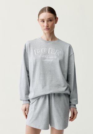 Nainen pukeutuneena vaaleanharmaaseen ylisuurikokoiseen collegepaitaan, jossa lukee "BORN Athleisure culture", sekä siihen sopiviin shortseihin, seisoo yksinkertaisen vaalean taustan edessä.