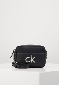 Sort læder crossover taske med et sølvfarvet "ck" logo, lynlåslukning og kædebelagt rem. Kompakt rektangulært design, glat tekstur.