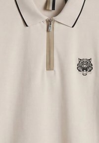 Polo beige con ribete negro en el cuello, cremallera frontal y un logo de cabeza de tigre negra en el pecho izquierdo.