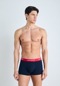 Boxers bleu marine avec une ceinture rouge Lacoste. Tissu doux et extensible ; coupe ajustée ; design minimaliste sans motifs ni embellissements.