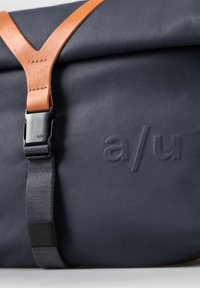 Borsa blu navy con una tracolla in pelle, caratterizzata da una superficie testurizzata e logo impresso. Fibbia regolabile e accenti a contrasto.
