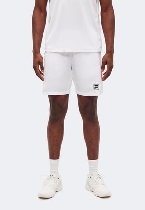 Mężczyzna w białych szortach sportowych, białych sneakersach i białych skarpetkach stoi z rękami swobodnie opuszczonymi wzdłuż ciała na jasnym tle.