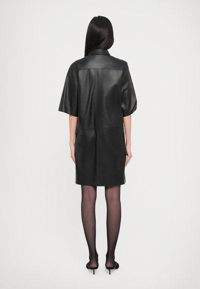 Γυναίκα με μακριά μαύρα μαλλιά που στέκεται με την πλάτη στραμμένη, φορώντας ένα μαύρο δερμάτινο φόρεμα μέχρι το γόνατο, διαφανείς μαύρες καλσόν και μαύρες γατούλες.