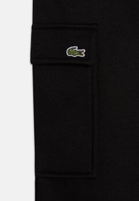 Un short en coton noir comporte une poche latérale et un logo brodé vert. La texture est lisse avec une finition légèrement extensible.