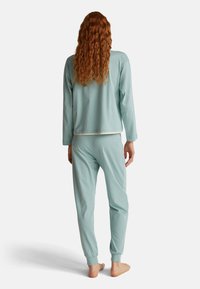 Persona con largo cabello rojo que lleva una camiseta de manga larga azul claro y pantalones jogger a juego, de pie descalza de espaldas sobre un fondo blanco.