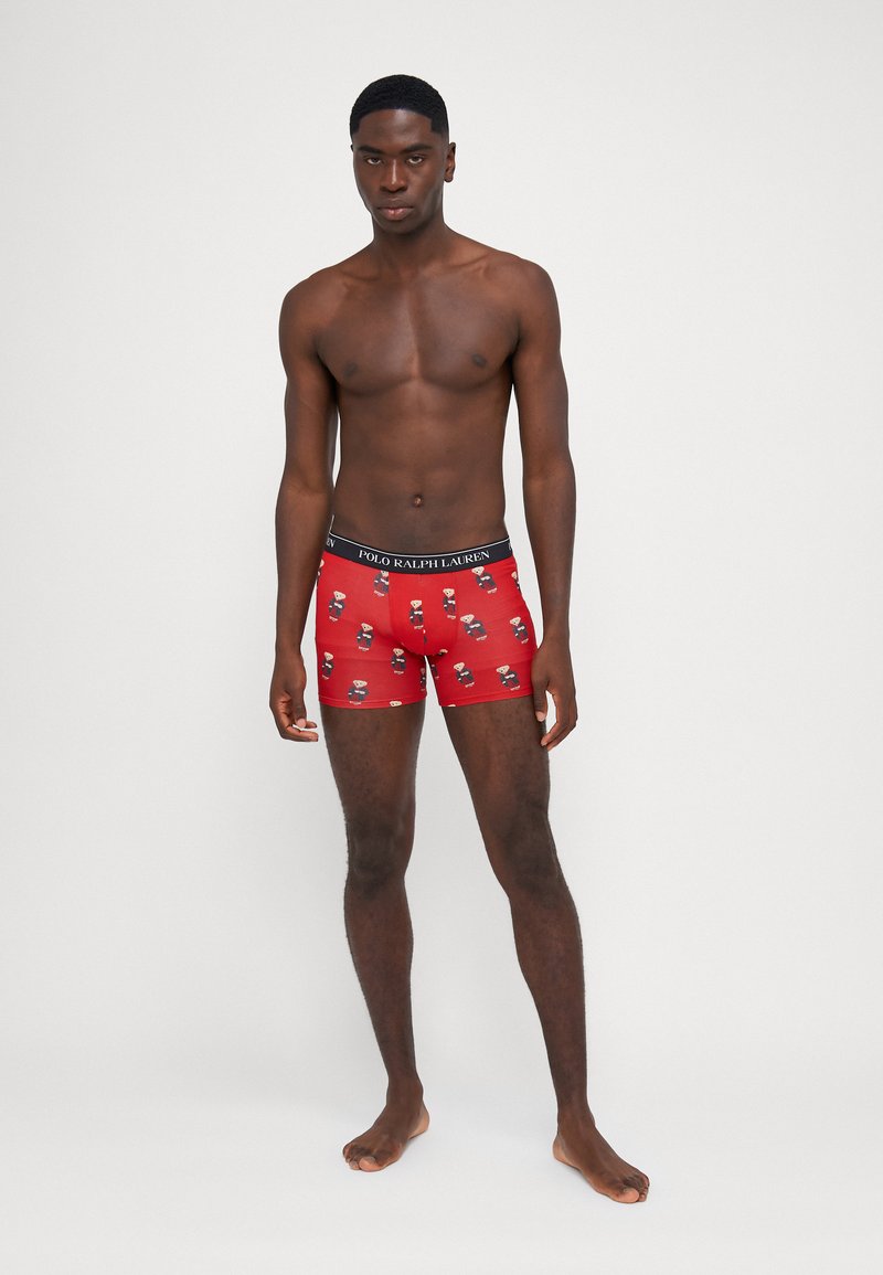 Shorts de boxeur rouges avec un motif de teddy bears en dessin animé. Fabriqués en tissu doux avec une ceinture noire affichant le nom de la marque.