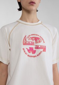 Valkoinen puuvillapaita, jossa on raglan-hihat. Paidassa on kiiltävä vaaleanpunainen logo-printti ja kontrastiväriset keltaiset ompeleet kauluksen ja hihansuiden ympärillä.