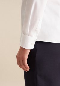 Polsino di camicia bianca a maniche lunghe con dettaglio a bottone, tessuto liscio, design su misura, abbinato a pantaloni blu scuro, in primo piano sul braccio.