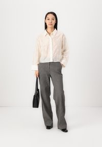 Lace white button-up shirt with long sleeves, paired with gray pinstriped wide-leg pants. Holds a black handbag, wearing black shoes.