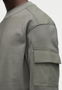 Φόρμα σε χρώμα τέφρας με ριμπ λαιμόκοψη, υφή υφάσματος και μεγάλη τσέπη-μπαλώματα στο αριστερό μανίκι. Διαθέτει ενισχυμένες ραφές στους ώμους.