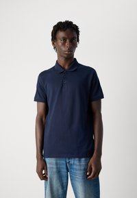 Fiatal férfi, rövid rasztafrizurával, sötétkék pólót és világoskék farmert viselve, egy sima fehér háttér előtt áll.