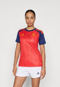 Woman wearing a red and blue Spain soccer jersey with the national emblem and white Adidas shorts, standing against a plain background.