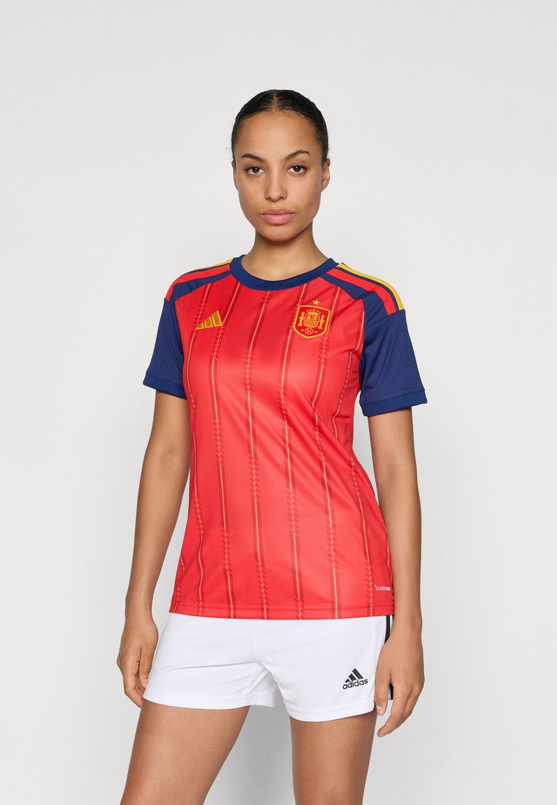 Woman wearing a red and blue Spain soccer jersey with the national emblem and white Adidas shorts, standing against a plain background.