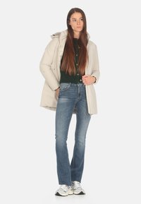 Giacca puffer grigio chiaro con cappuccio, indossata sopra un maglione di lana verde scuro e jeans a zampa blu, abbinata a sneakers argento e nere.