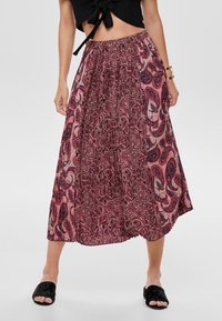 Una persona sta in piedi indossando un top nero a maniche corte e una gonna lunga fluida e a fantasia in tonalità di rosa e bordeaux, abbinata a sandali neri.