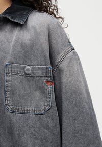Γκρι τζιν μπουφάν με τσέπη στο στήθος που διαθέτει μπλε ραφές και ένα μικρό κόκκινο κεντητό λογότυπο στην επάνω αριστερή πλευρά.