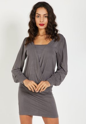 Mujer con cabello rizado oscuro, vestida con un vestido envolvente de manga larga gris, de pie contra un fondo blanco liso.