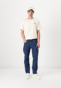 Világos bézs T-shirt kerékpár grafikával, kék egyenes lábú farmernadrág és fehér sportcipő. Egy világos sapka teszi teljessé a laza megjelenést.
