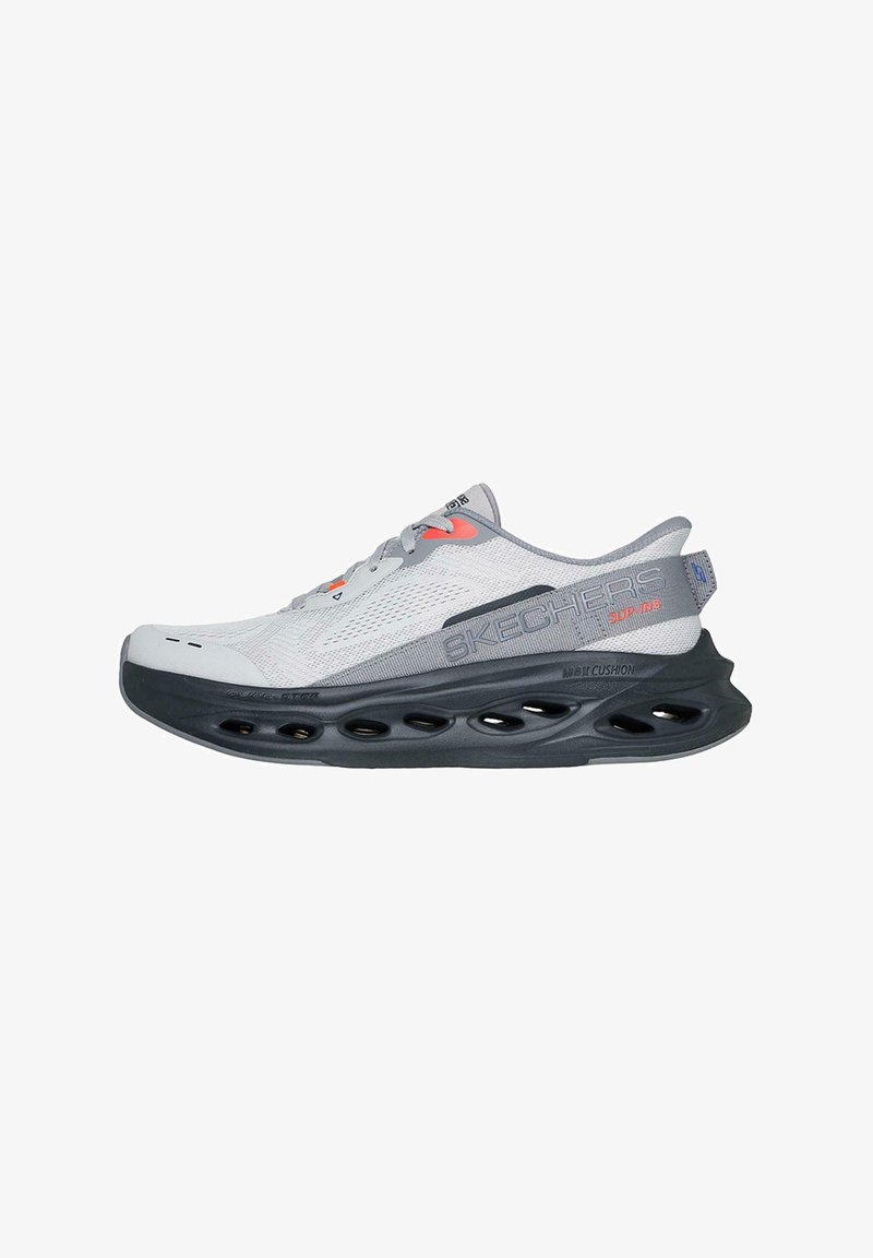 Zapatilla atlética gris con parte superior de malla, entresuela negra con recortes y detalles en naranja. Diseñada para comodidad y facilidad al ponérsela.