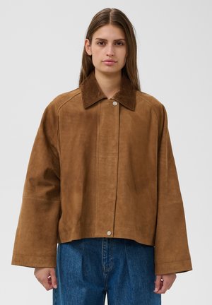 Femme portant une veste en daim marron oversize avec un col en velours côtelé et un jean bleu, debout devant un fond gris uni.