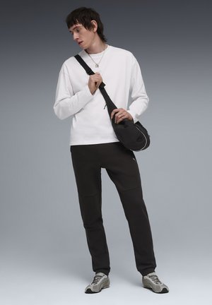 Camisa blanca de manga larga, pantalones jogger negros con detalles de parches, zapatillas grises y un pequeño bolso cruzado negro con detalles blancos.