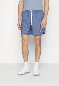 Blå badshorts med dragsko i midjan, sidofickor och en liten vit logotyp. Bärs med vita träning skor och strumpor.