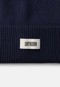 Námornícky pletený čiapka s ryhovaným vzorom, ktorá má bielu etiketu s nápisom "DRYKORN" v čiernom písmene. Tkanina je štruktúrovaná a pružná pre pohodlné nosenie.