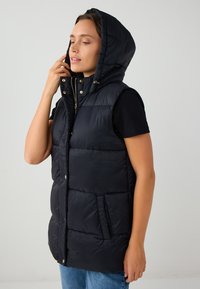 Gilet imbottito nero con cappuccio, dotato di zip frontale, bottoni a scatto e due tasche laterali. Texture liscia con design trapuntato.