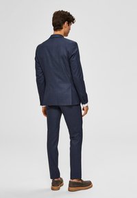 Marineblå dress med en strukturert blazer og slim-fit bukser, med subtil tekstur og knappe-ermer. Brune sko fullfører antrekket.