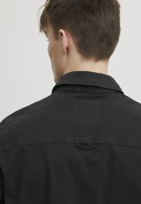 Black shirt featuring a classic collar, visible stitching, and a back loop detail for hanging. The material appears smooth and textured.