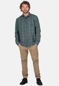 Mann trägt ein karierter Hemd in Grün, beigetöne Hosen und schwarze Schuhe, steht mit den Händen in den Taschen vor einem weißen Hintergrund.