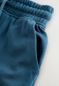 Pantalons de jogging teal en tissu texturé, avec une taille élastique et une poche latérale. Le matériau semble doux et légèrement extensible.