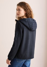 Veste marine avec une capuche matelassée en couches, des poignets côtelés et une poche latérale en tissu contrasté. Texture douce, coupe décontractée.