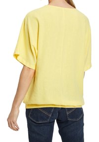 Żółta bluzka z krótkim rękawem o luźnym kroju, okrągłym dekolcie i miękkim, teksturowanym materiale. Noszona z ciemnoniebieskimi dżinsami, widok z tyłu.