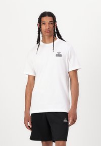 White cotton t-shirt with a round neck and short sleeves, featuring a gray logo on the left chest. Worn with black shorts.